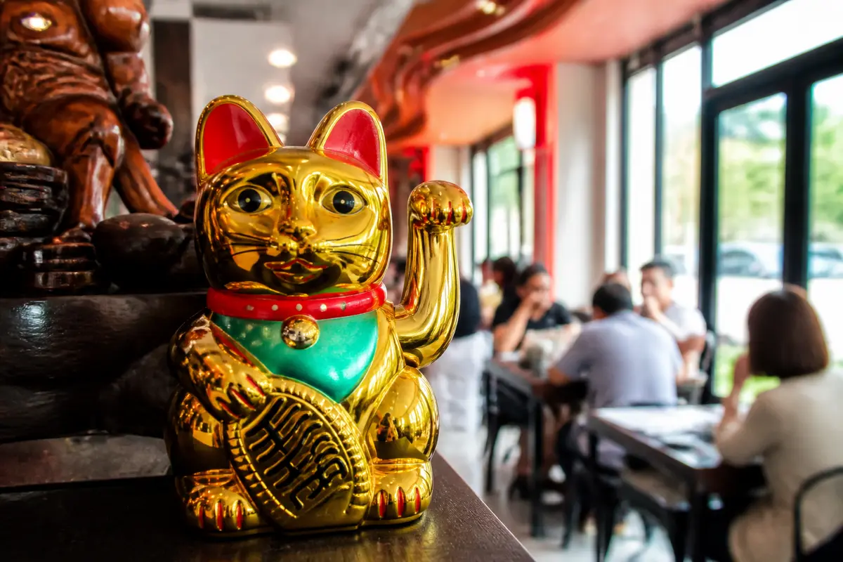 Chat porte-bonheur doré sur étagère, restaurant animé à Saint-Brice-sous-Forêt en arrière-plan